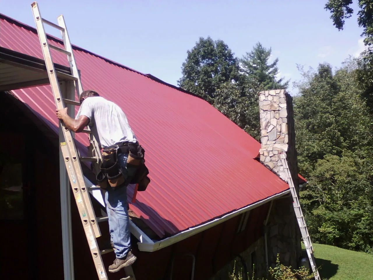 Roofer on a ladder beside a red metal roof and stone chimney — Soffit & Fascia Repair by Roofing Brothers in Ozark
